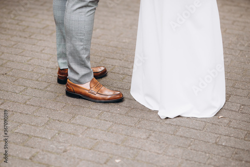 Bride and groom stand against each other. Groom with leather brown lacquered shoes, grey suit pants and the bride with white dress. Concrete pavement. Love, outdoor wedding,celebration and good mood.