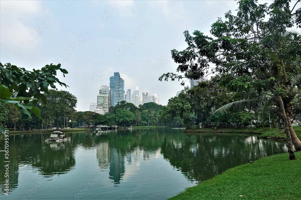 Fototapeta premium Summer day in the Bangkok park. Boats float on the quiet lake. Green grass and palm trees on the shore. Silhouettes of skyscrapers against the clear sky. Reflection in water. Thailand. Lumpini park.