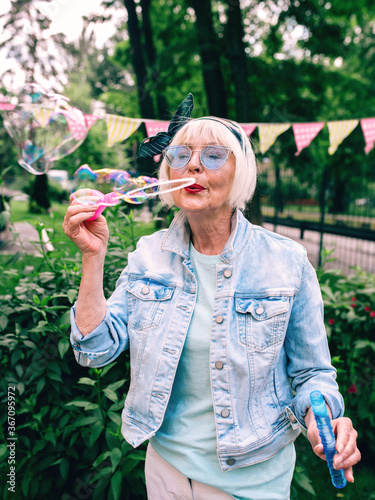 senior (old) stylish woman with gray hair and in blue glasses and jeans jacket blowing bubbles outdoors. Holidays, party, anti age, fun concept