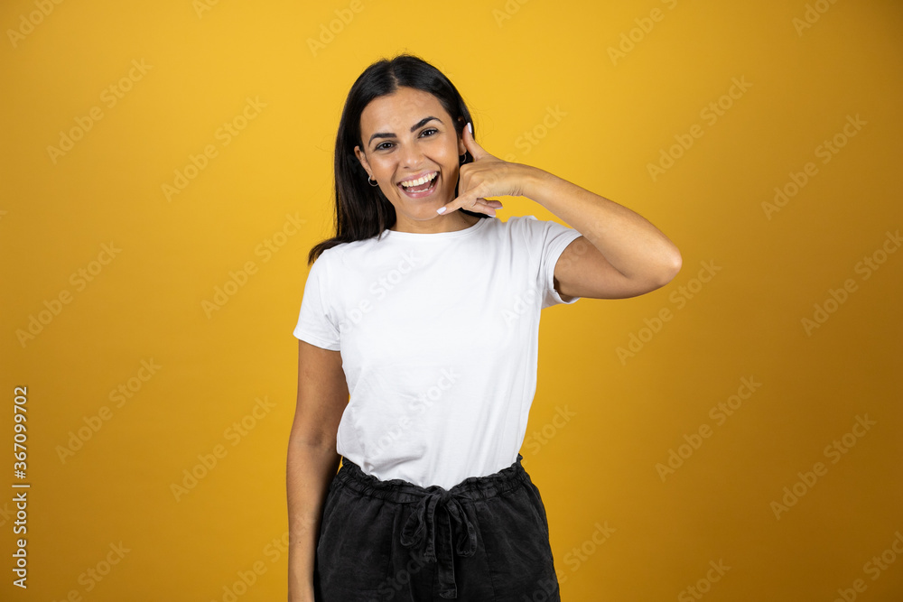 Young beautiful woman smiling and putting her hand next to her ear like it was a telephone. Wearing casual white t-shirt on yellow background Young beautiful woman smiling and putting her hand next to her ear like it was a telephone. Wearing casual white t-shirt on yellow background