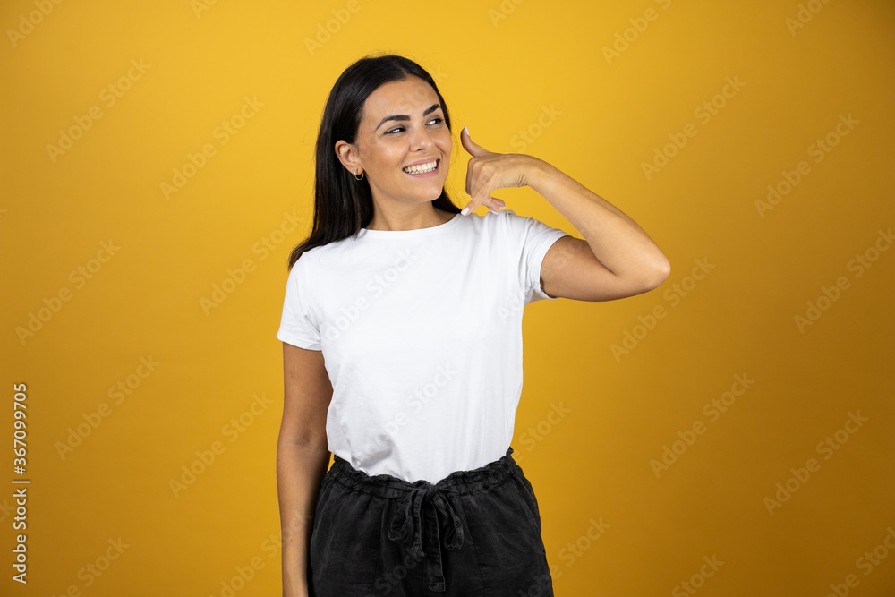 Young beautiful woman smiling and putting her hand next to her ear like it was a telephone. Wearing casual white t-shirt on yellow background Young beautiful woman smiling and putting her hand next to her ear like it was a telephone. Wearing casual white t-shirt on yellow background