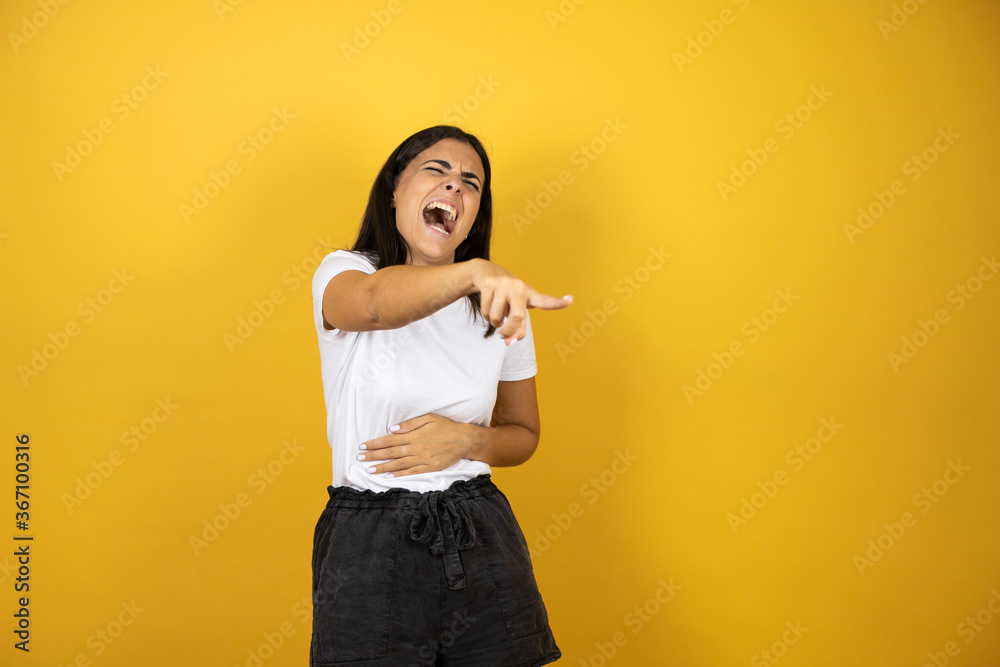 Young beautiful woman over yellow background laughing at you, pointing finger to the camera with hand over body, shame expression Young beautiful woman over yellow background laughing at you, pointing finger to the camera with hand over body, shame expression
