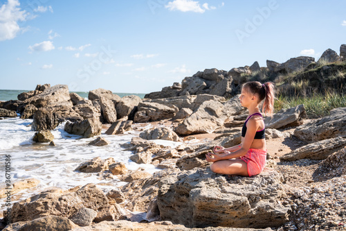 A girl sits in a lotus position, meditates face to the sea on a stone, side view. in front of her is the sea,
