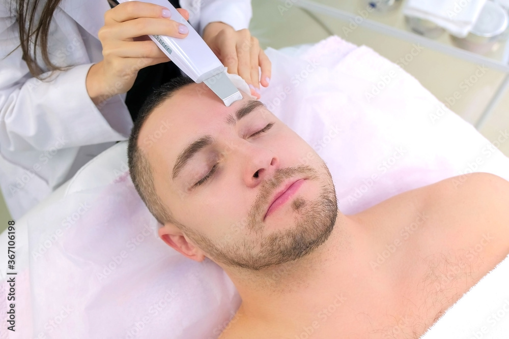 Fototapeta premium Cosmetologist woman is making ultrasonic face cleaning procedure for young man in clinic. Closeup client portrait and beautician's hands. Procedure in cosmetology salon. Beauty industry concept.