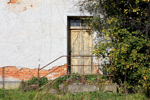 Wooden dilapidated entrance doors to abandoned ruins of suburban family house with broken steps and rusted metal fence surrounded with uncut overgrown grass and trees