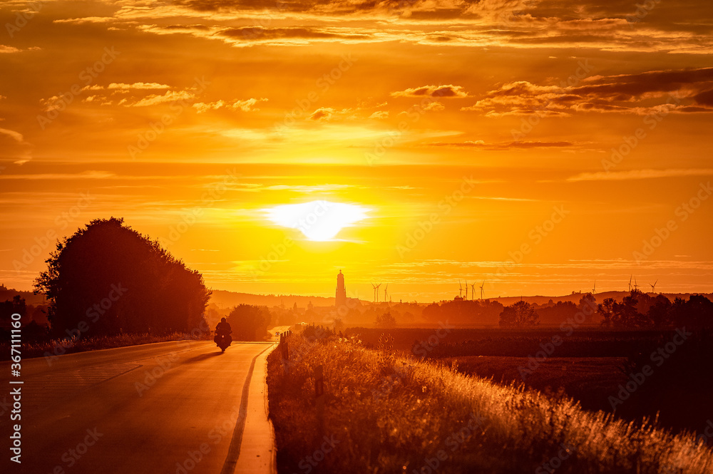Beautiful scene of a main road in the sunset ending in the horizont with Nördlingen in Bavaria Germany. A motorbike is driving down the road towards the suns.