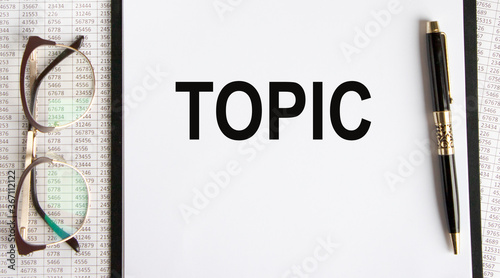 On the paper tablet is the inscription TOPIC, next to glasses and a pen, all against the background of accounting.