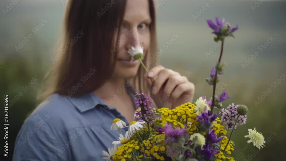 A blonde woman in a denim dress with wild flowers walks through a summer field with high green grass on a rural hill. A blonde woman in a denim dress with wild flowers walks through a summer field with high green grass on a rural hill.