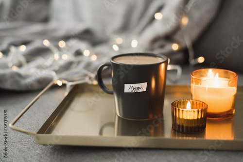 Hygge concept: hot coffee in black mug served on a metal tray with two burning candles, decorative lights as background. Cozy atmosphere