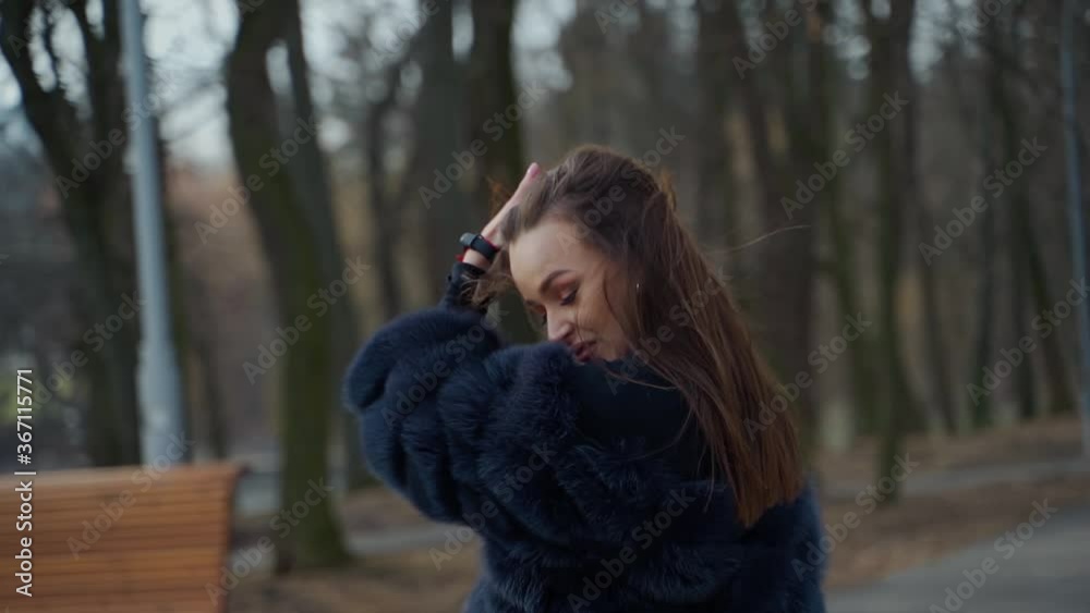 Attractive girl in the city park. Stylish woman in fur coat turning around and looking on camera outdoors. Slow motion.