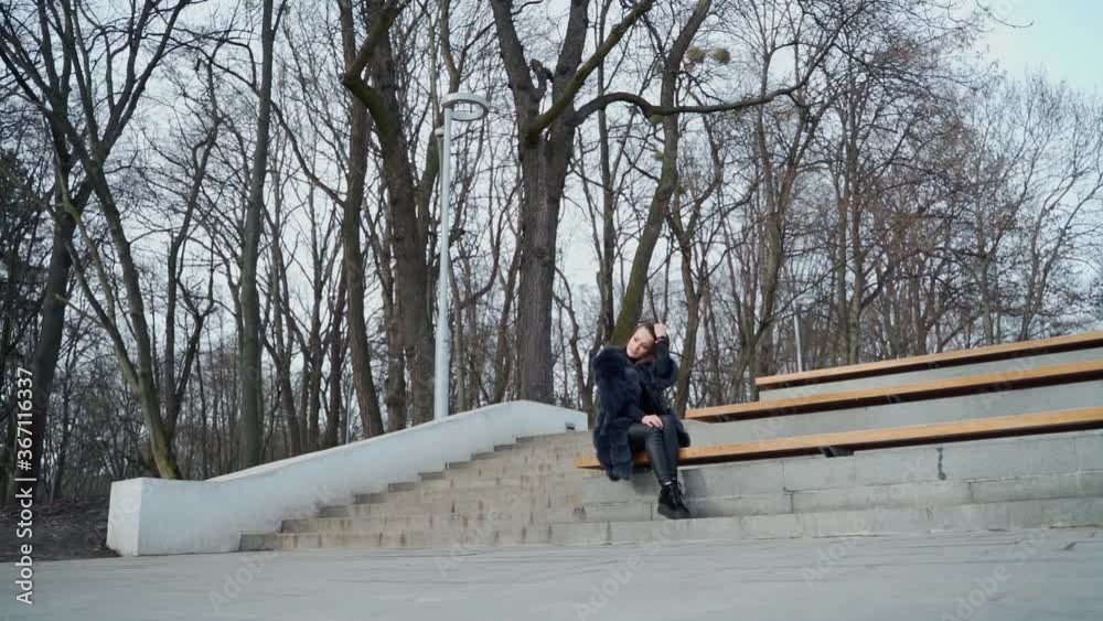 Girl sits on bench in the park. Beautiful young woman in fluffy coat sitting alone outdoors on the leafless trees background. View from below. Girl sits on bench in the park. Beautiful young woman in fluffy coat sitting alone outdoors on the leafless trees background. View from below.