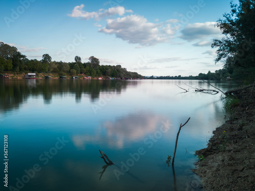 Landscape of the Sava river with fishing huts by the water and banks overgrown with dense forest during a sunny day. A few cumulus clouds in the sky and a beautiful natural setting.