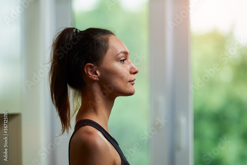 A side view photo of a sporty slim girl in a black workout tight suit is relaxing before training near the window at home.