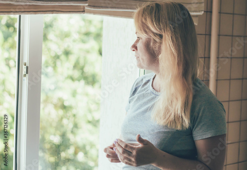  Woman at home looks thoughtfully out the window and drinks coffee