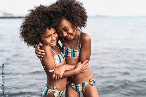 Happy sisters embracing inside sea water during summer time - Afro kids having fun playing on the beach - Family love and travel vacation lifestyle concept