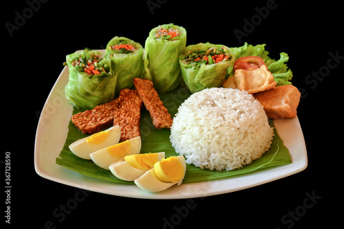 Traditional indonesian meal made of rice, vegetable spring rolls, fried tempeh and hard boiled eggs, isolated on black background.
