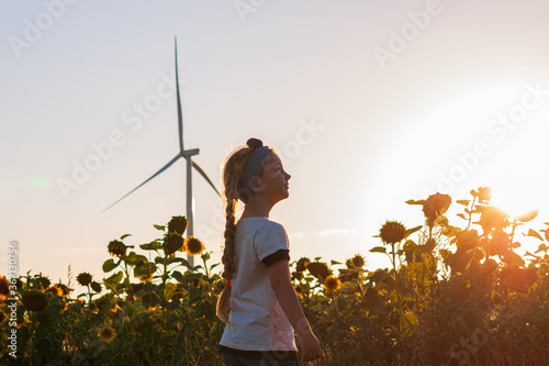 Cute girl in white t-shirt smelling sunflower in sunset field with wind turbines farm on background. Child with long braid hair on countryside landscape with yellow flowers. Farming concept wallpaper.