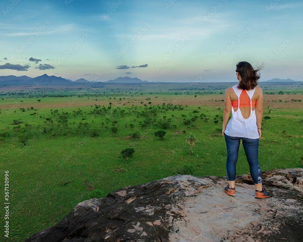 Fototapeta premium Young woman looking into the horizon of the beautiful landscapes of Kidepo Valley National Park, Uganda, Africa. The woman rests on top of a kopje after a nice hike.