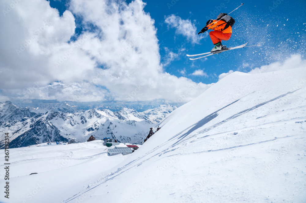 A male skier in an orange suit flies in the air after jumping from a snow sweep high in the Caucasian mountains on a sunny day amid snow-capped mountains of blue sky and white clouds
