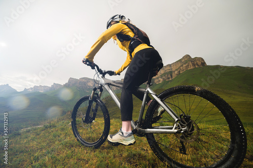 Attractive red-haired girl in a helmet and with a backpack sits on a mountain bike in the mountains against the backdrop of rocks and cloudy sky