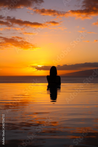 a Girl stands with her back to the pool  against the background of an orange sunset and the ocean.