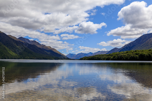 The calm waters of South Mavora Lake with forested mountains down each side and mountains in the distance. White Fluffy clouds on a blue sky.