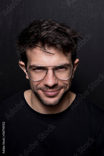 Retrato de joven apuesto sonriendo, usa lentes y piercing en la oreja, remera negra y fondo negro photo