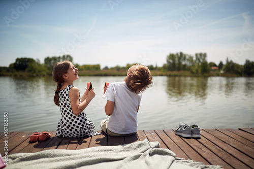 cute minor brother and sister laughing at riverside, sitting by the river eating watermelon