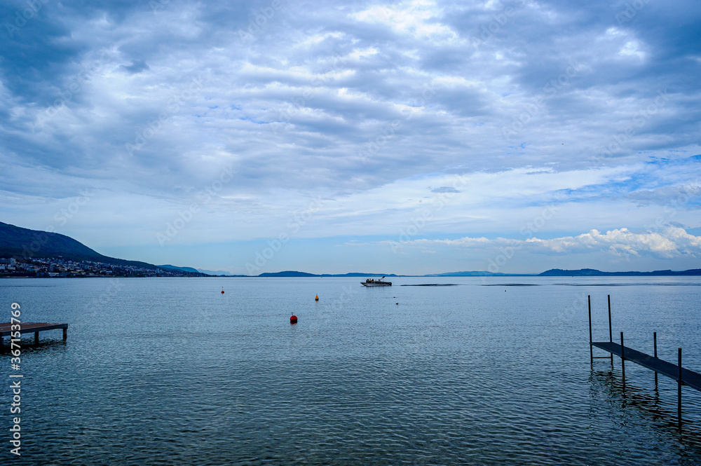 Photo taken with a wide-angle lens. View of Lake Neuchâtel at blue time. The perspective makes two footbridges converge towards a boat passing in the background.  Cloudy sky over the lake.