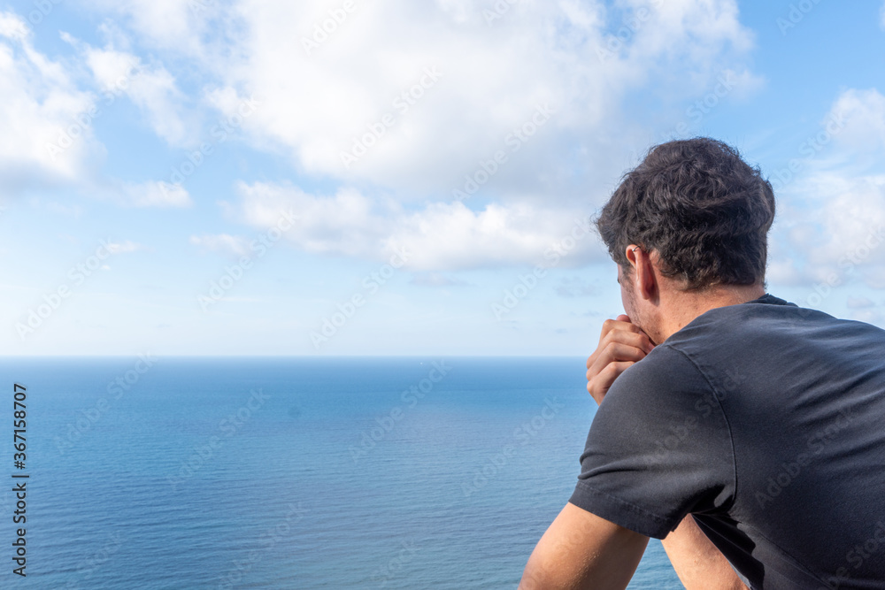 Close-up of young man on his back leaning against a wooden railing, looking at a sunny seascape with white clouds, horizontal