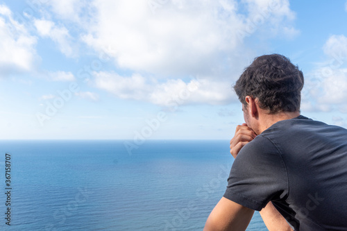 Close-up of young man on his back leaning against a wooden railing, looking at a sunny seascape with white clouds, horizontal