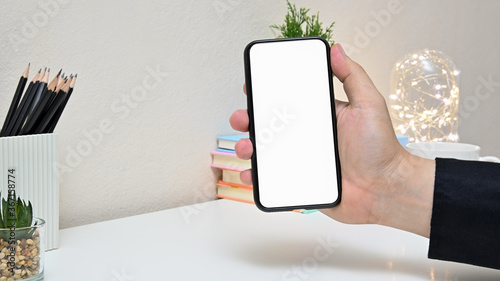 Front view of man’s hand holding smartphone on white table in home office with lightbulb, cactus and blank screen smartphone. concept work from home.