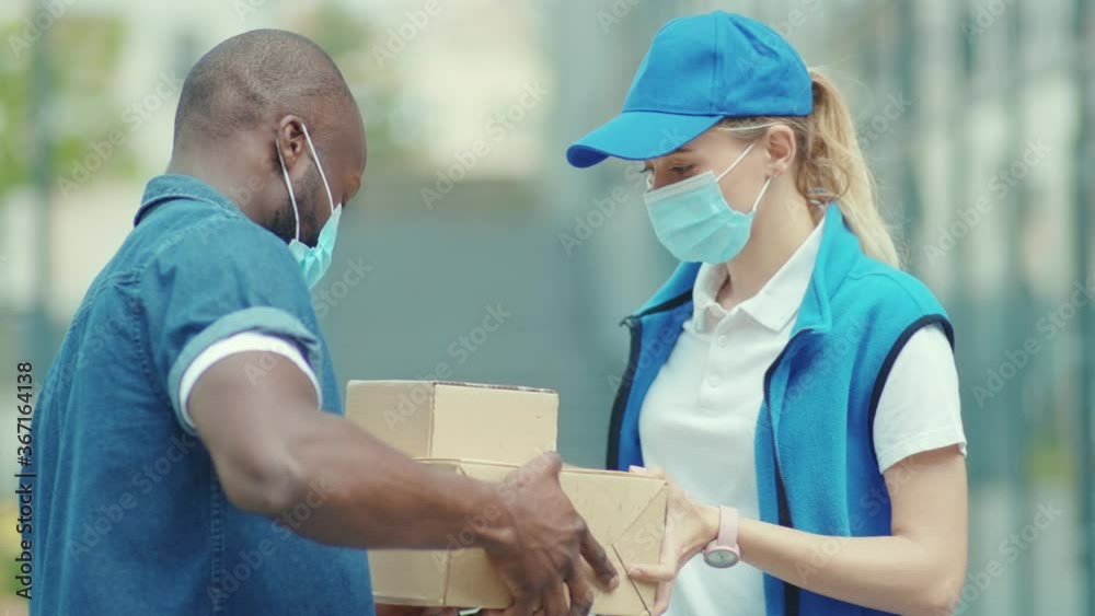Portrait shot of young pretty female courier in blue hat and in medical mask give carton parcel to african american man worker outdoors slow motion Portrait shot of young pretty female courier in blue hat and in medical mask give carton parcel to african american man worker outdoors slow motion