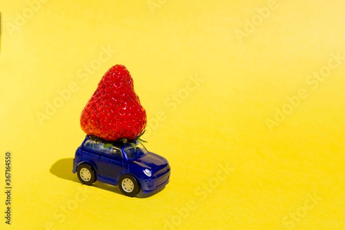 Close-up of small blue car carrying huge ripe strawberry. Food delivery and dessert production concept. Sweet seasonal present ..