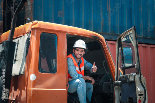 Handsome foreman worker wearing safety equipment, giving thumb up and smiling in truck at logistic shipping cargo containers yard