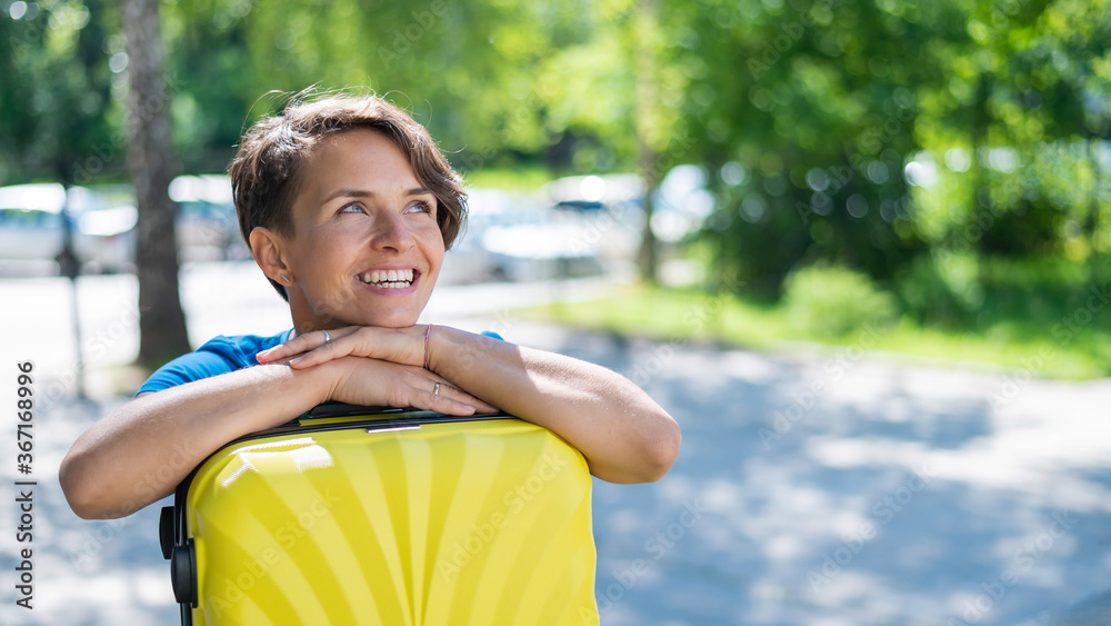 Close-up portrait of a beautiful short-haired Caucasian young woman with a yellow suitcase outdoors. The girl with a perfect smile dreamily leaned her head against a large luggage bag. Close-up portrait of a beautiful short-haired Caucasian young woman with a yellow suitcase outdoors. The girl with a perfect smile dreamily leaned her head against a large luggage bag.