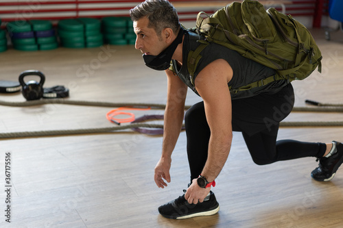Motivated instructor kneeling down and ties his shoestrings with backpack and training mask. Starting training. For boot camp and gym concept.