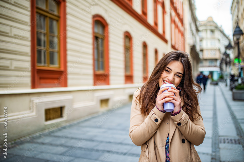 Beautiful woman walking outdoors on a sunny day. Beautiful young woman holding coffee cup and smiling while walking along the street. Portrait of a stylish girl with takeaway coffee