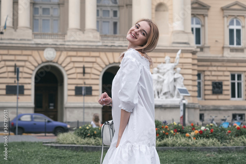 Gorgeous young model girl with perfect blonde hair looking at camera posing in the old city wearing white dress.