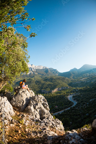 A woman with a backpack sits on top of a mountain and admires the beauty of a mountain valley.