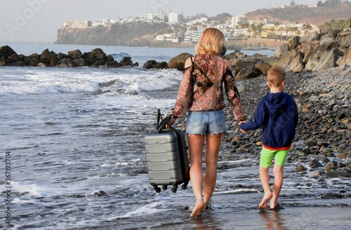 young slender woman walks with a suitcase and a boy along the ocean