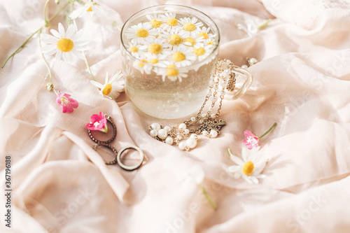 Daisy flowers in water in glass cup on background of soft beige fabric with wildflowers and jewelry. Tender floral aesthetic. Creative summer image with space for text. Bohemian mood