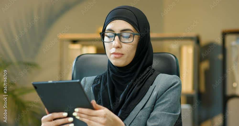Portrait of Arab beautiful woman in black hijab sitting in office, tapping and scrolling on tablet device screen. Muslim happy businesswoman in headscarf smiling to camera. Arab female in glasses. Portrait of Arab beautiful woman in black hijab sitting in office, tapping and scrolling on tablet device screen. Muslim happy businesswoman in headscarf smiling to camera. Arab female in glasses.