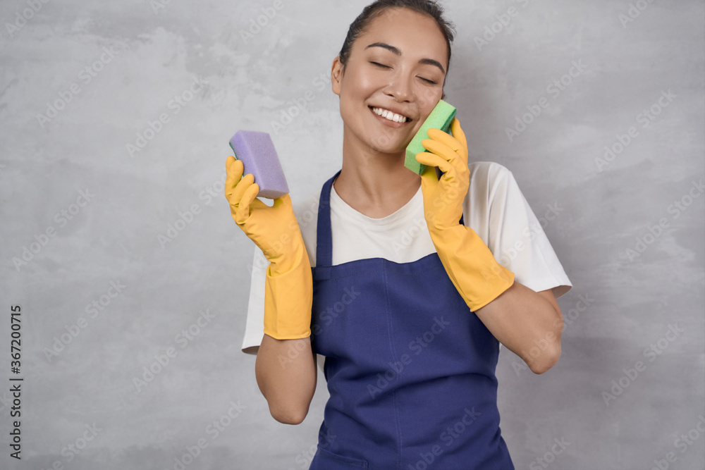 Washing dishes with fun. Happy young cleaning lady in yellow rubber gloves playing with kitchen sponges and smiling while standing against grey wall Washing dishes with fun. Happy young cleaning lady in yellow rubber gloves playing with kitchen sponges and smiling while standing against grey wall