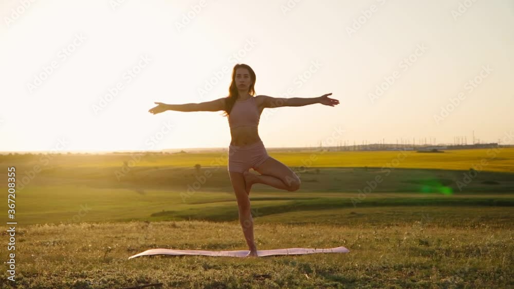 Young woman is practicing yoga outdoors in park evening on background beautiful field and sunset. Fitness in nature. Stretching health and wellness. Sport Motivation. Slow motion Young woman is practicing yoga outdoors in park evening on background beautiful field and sunset. Fitness in nature. Stretching health and wellness. Sport Motivation. Slow motion