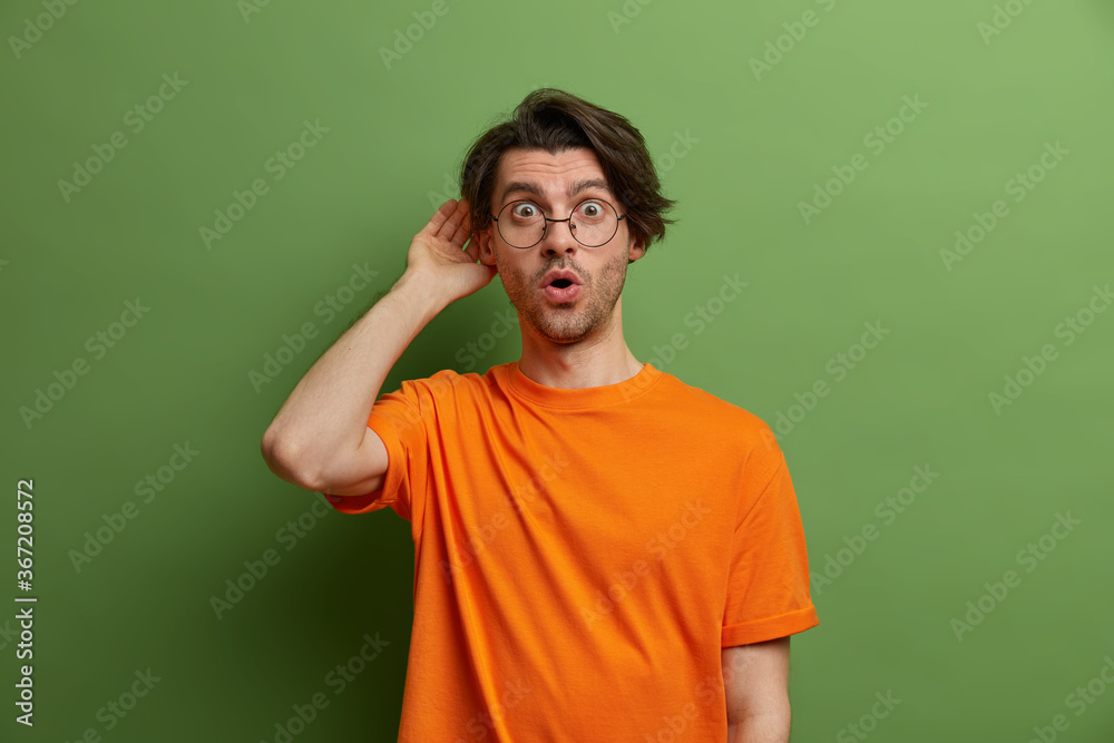 Stunned emotional guy eavesdrops information, keeps hand near ear, shocked to hear something unexpected, surprised by gossips, wears glasses and orange t shirt, stands against green background Stunned emotional guy eavesdrops information, keeps hand near ear, shocked to hear something unexpected, surprised by gossips, wears glasses and orange t shirt, stands against green background