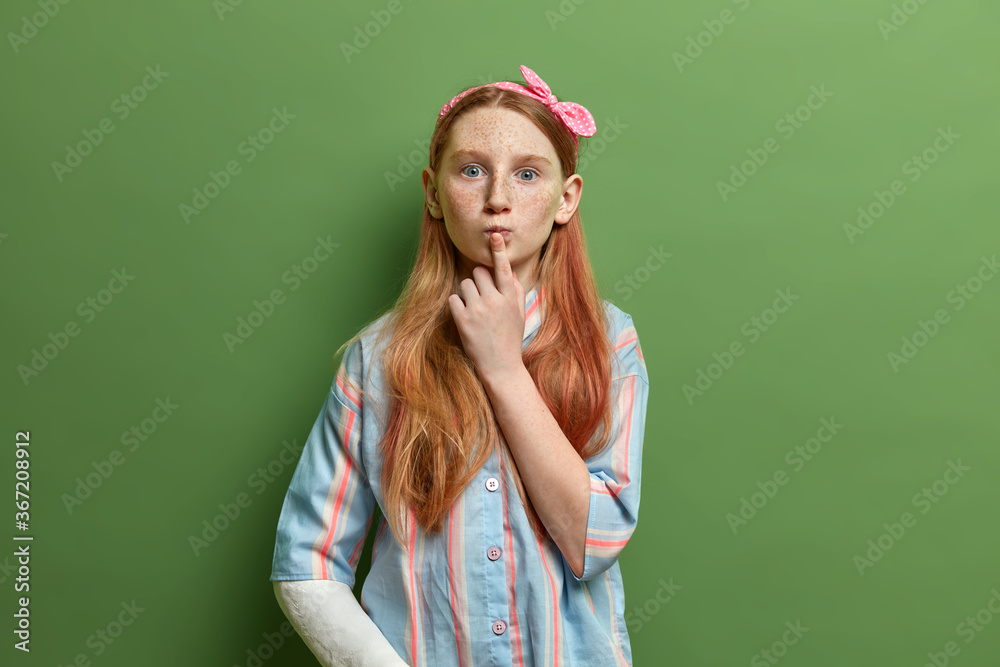 Beautiful surprised freckled girl keeps finger on rounded lips, has curious expression, listens something with interest, wears headband and shirt, has broken arm, isolated on green studio wall Beautiful surprised freckled girl keeps finger on rounded lips, has curious expression, listens something with interest, wears headband and shirt, has broken arm, isolated on green studio wall