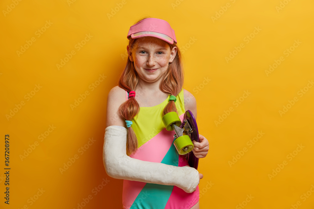 Beautiful carefree girl with two pony tails, glad after riding skateboard, wears cast on broken arm, dressed in bathing suit and cap, spends free time in skatepark, isolated on yellow background Beautiful carefree girl with two pony tails, glad after riding skateboard, wears cast on broken arm, dressed in bathing suit and cap, spends free time in skatepark, isolated on yellow background