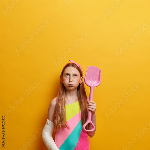 Fotografija Small gloomy girl with long red hair, blows cheeks and looks upwards, plays on s
