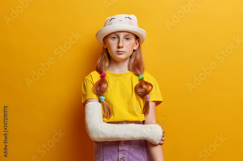 Calm serious small girl looks directly at camera, has long ginger hair and freckled skin, wears fashionable summer outfit, poses with hand in cast, recovers after accident, isolated over yellow wall